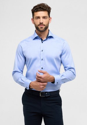 Hellblaues Langarmhemd mit glatter Textur, taillierter Passform, gemustertem Innenkragen und dunklen Knöpfen. Kombiniert mit navyblauen Hosen.