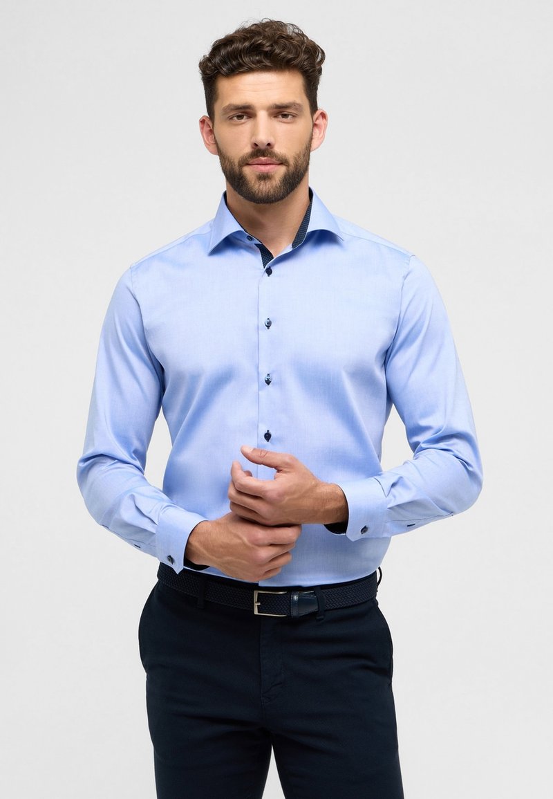 Camicia azzurra a maniche lunghe con una texture liscia, vestibilità aderente, colletto interno a fantasia e bottoni scuri. Abbinata a pantaloni blu navy.