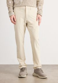 Persoon in beige rechte broek, taupe sneakers en een beige gebreid vest staat op een grijze vloer tegen een effen witte muur.