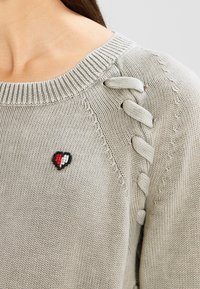 Gros plan d'un pull en tricot beige avec col côtelé, détails de laçage sur l'épaule et petit cœur brodé en rouge, blanc et noir sur la poitrine.