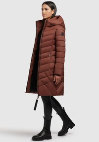 Brauner gepolsterter langer Mantel mit Kapuze, der einen Reißverschluss und ein gestepptes Design aufweist. Kombiniert mit schwarzen Lederleggings und Ankle Boots.