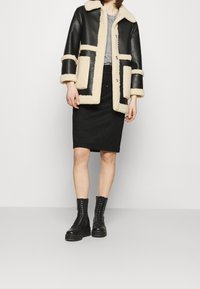 Veste en cuir noir avec finitions en shearling beige, présentant un col, deux grandes poches avant et une silhouette carrée, portée avec une jupe noire et des bottes.