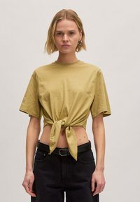 Olijfgroen T-shirt met korte mouwen en een geknoopte voorkant, voorzien van een ronde halslijn en zachte textuur, gecombineerd met zwarte high-waisted jeans.
