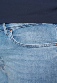 Světle modré džíny s klasickým pětikapsovým designem, viditelným prošíváním a měděnou nýtkou na přední kapse. Hladká denimová textura.