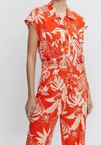 Top corto floreale rosso con maniche corte e chiusura a bottoni, abbinato a pantaloni a vita alta con un motivo di foglie tropicali.