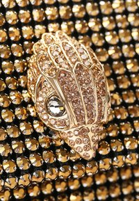 Accessorio decorativo color oro a forma di mano con molteplici abbellimenti simili a gemme, su uno sfondo testurizzato nero e oro.