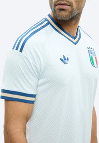 Muž nosící bílý italský fotbalový dres s modrým a béžovým lemováním, logem Adidas a italským znakem se čtyřmi hvězdami na hrudi.