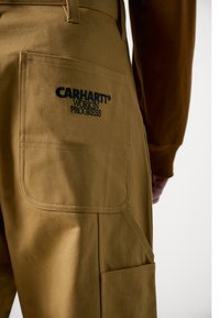 Braune Arbeitshosen aus robustem Stoff, mit einer auffälligen Gesäßtasche, auf der in Dunkelblau "CARHARTT WORK IN PROGRESS" gestickt ist.