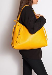 Geel lederen handtas met een textuurfinish, verstelbare schouderriem en een decoratieve charm in de vorm van een beer. Ruimtelijk, slungelig ontwerp.