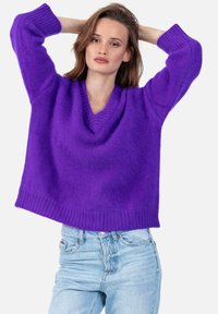 Pull-over ample à col en V en tissu violet vif et doux. Comprend des poignets et un ourlet côtelés, porté sur un jean slim bleu clair.