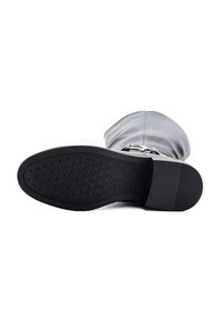 Semelle noire d'une botte en cuir grise, hauteur genou, avec un détail en boucle, présentée à plat sur un fond blanc.