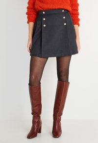 Falda A-line de color gris oscuro con acentos de botones dorados, combinada con medias negras y botas de cuero marrón de tacón alto hasta la rodilla. Suéter de punto texturizado en color naranja.