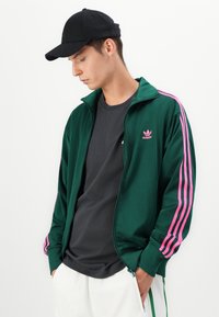 Grønn treningjakke med rosa striper, med glidelås og en liten rosa logo. Bæres over en mørk t-skjorte og kombinert med en svart caps.
