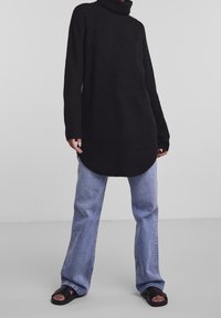 Svart stickad polotröja med långa ärmar, tillsammans med ljusblå vida jeans och svarta sandaler, med en rundad nederkant.