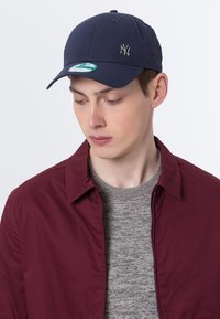 Marineblaue Baseballmütze mit einem Logo vorne, gebogenem Schirm und verstellbarem Riemen hinten. Tragen mit einer burgunderfarbenen Jacke und einem grauen Pullover.