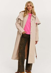 Žena v béžovém trenčkotě, jasně růžovém svetru a kalhotách s leopardím potiskem, stojící s jednou rukou v kapse na bílém pozadí.