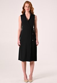 Zwarte mouwloze wikkeljurk met kraag, gestructureerde stof, zijdetail met strik en geplooide roksectie. Draagt beige hakken.