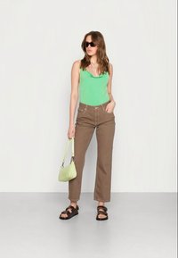Top verde sem mangas, jeans castanhos de cintura alta, sandálias castanhas escuras e uma pequena bolsa verde. Modelo permanece em um fundo neutro.