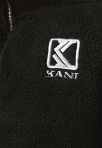 Jachetă din fleece neagră cu o suprafață texturată, logo brodat alb și fermoar în față. Accentuată de un design logo pătrat.