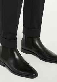 Black leather Chelsea boots with a sleek design, featuring elastic side panels and pointed toes, paired with tailored black trousers.