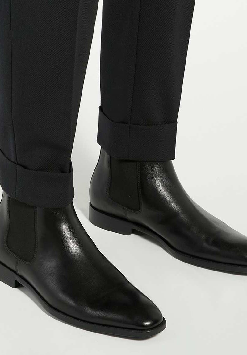 Black leather Chelsea boots with a sleek design, featuring elastic side panels and pointed toes, paired with tailored black trousers.