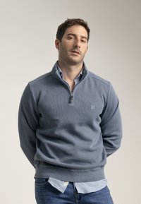Suéter gris de punto con cuello con cremallera, textura acanalada y dobladillo elástico. Llevado sobre una camisa de color azul claro con cuello, que presenta un pequeño parche de logo.