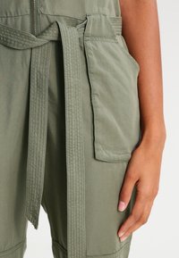 Prawa ręka spoczywa obok zielonego kombinezonu roboczego z przeszytym paskiem i dużą boczną kieszenią z klapką.