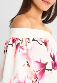 Femme portant une robe blanche à épaules dénudées avec des motifs floraux roses et violets, montrant son épaule nue et une partie de son visage souriant.