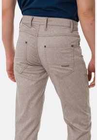 Beige strukturierte Hose mit Gesäßtaschen, die ein dezentes Muster aufweist. Metallknopfakzente und ein Etikett an der rechten Tasche.