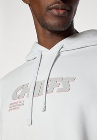 Bela kapuca z logotipom "CHIEFS" v sivi in rdeči barvi. Mehka tekstura z nastavljivo vrvico in žepom za roke.