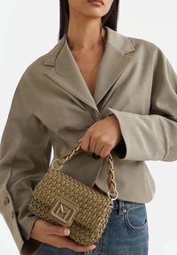 Borsa intrecciata beige con tracolla a catena dorata, caratterizzata da un logo quadrato sul davanti, tenuta da una persona con un blazer marrone chiaro.