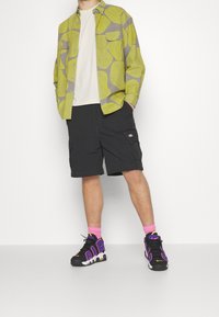 Chemise à motifs verts et gris portée sur un t-shirt blanc, shorts cargo noirs, chaussettes roses et baskets noires avec des accents violets.
