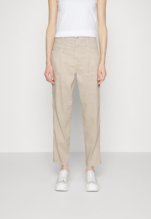 Frau trägt beige hoch taillierte Hose mit Falten und Vordertaschen, kombiniert mit weißen Sneakern und weißem ärmellosem Oberteil.