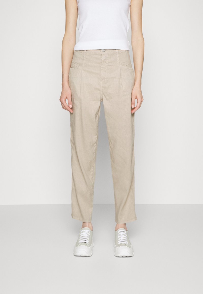 Femme portant un pantalon beige taille haute avec plis et poches avant, associé à des baskets blanches et un haut sans manches blanc.
