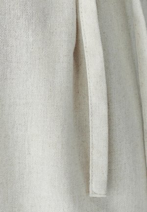 Detailní záběr na béžovou hrubou tkaninu s vroubkovaným obdélníkovým páskem nebo zavazováním visícím svisle.