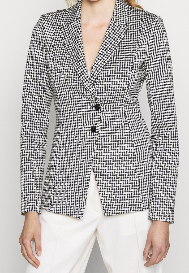 Femme portant un blazer cintré en pied-de-poule noir et blanc avec deux boutons noirs, assorti à un pantalon blanc sur un fond uni.