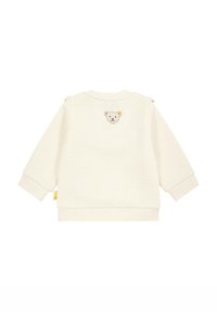 Sweatshirt couleur crème en tissu texturé, présentant une broderie de visage d'ours au dos et des fermetures pression sur l'épaule.