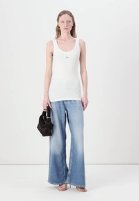 Top de tirantes blanco acanalado con un pequeño logo, combinado con unos vaqueros de pierna ancha de mezclilla de color azul claro y un pequeño bolso negro. La modelo está de pie frente a un fondo liso.