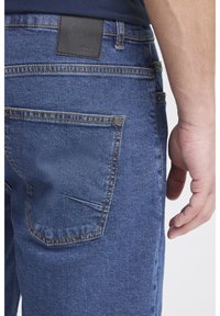 Modre kavbojke iz jeansa z zadnjim žepom, rumenim šivanjem in črno usnjeno značko na pasu. Gladek tekstura, raven kroj.