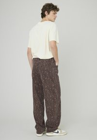 Pantalon marron à motifs avec une coupe décontractée, associé à une chemise à manches courtes de couleur claire. Tissu texturé avec des poches arrière visibles.