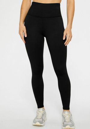 Woman wearing high-waisted black leggings and gray running shoes, standing with hands relaxed by sides on white background.