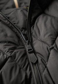 Sort quilted jakke med en robust lynlås, tekstureret stof og et logo på lynlåsgrebet. Glat indvendig foret synligt.