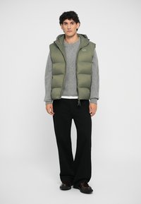 Olivový zelený puffový vest s kapucňou na svetlo šedej pletenej sveter a čierne široké nohavice. Tmavé topánky dopĺňajú outfit.