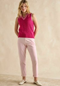 Rozā adīta veste ar V-veida kaklu un baltām svītrām ap apkakli, apvienota ar gaiši rozā izbrīvētiem biksēm un baltām loafer tipa kurpēm.