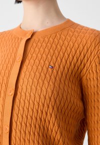 Gros plan sur une personne portant un cardigan orange en tricot torsadé avec des boutons, orné d'un petit logo rectangulaire rouge, blanc et bleu sur la poitrine.