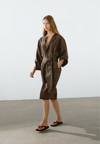 Robe portefeuille en lin brun avec un décolleté en V, des manches longues retroussées et des poches latérales. Associée à des sandales en plastique noir et rouge, debout sur un sol gris.
