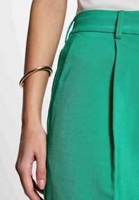 Pantaloni turchesi su misura con una finitura strutturata e pieghe frontali. Un bracciale dorato su una mano aggiunge un tocco metallico.