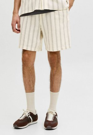 Mehe jalad, kes kannavad kreemikaid lühikesi pükse õhukeste mustade vertikaalsete triipudega, beežid sokid ja pruunid tossud valgete paelte ja taldadega.