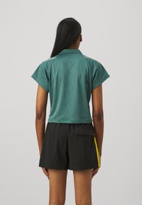 Groene gekorte polo met korte mouwen, voorzien van een subtiel patroon en witte schouderstrepen, gecombineerd met zwarte shorts met gele accenten.