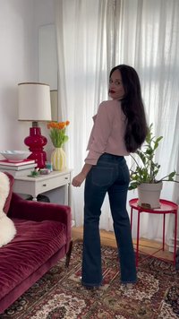 Kvinne med langt mørkt hår iført rosa topp og blå jeans står ved hvite gardiner i en koselig stue med planter og vintage møbler.